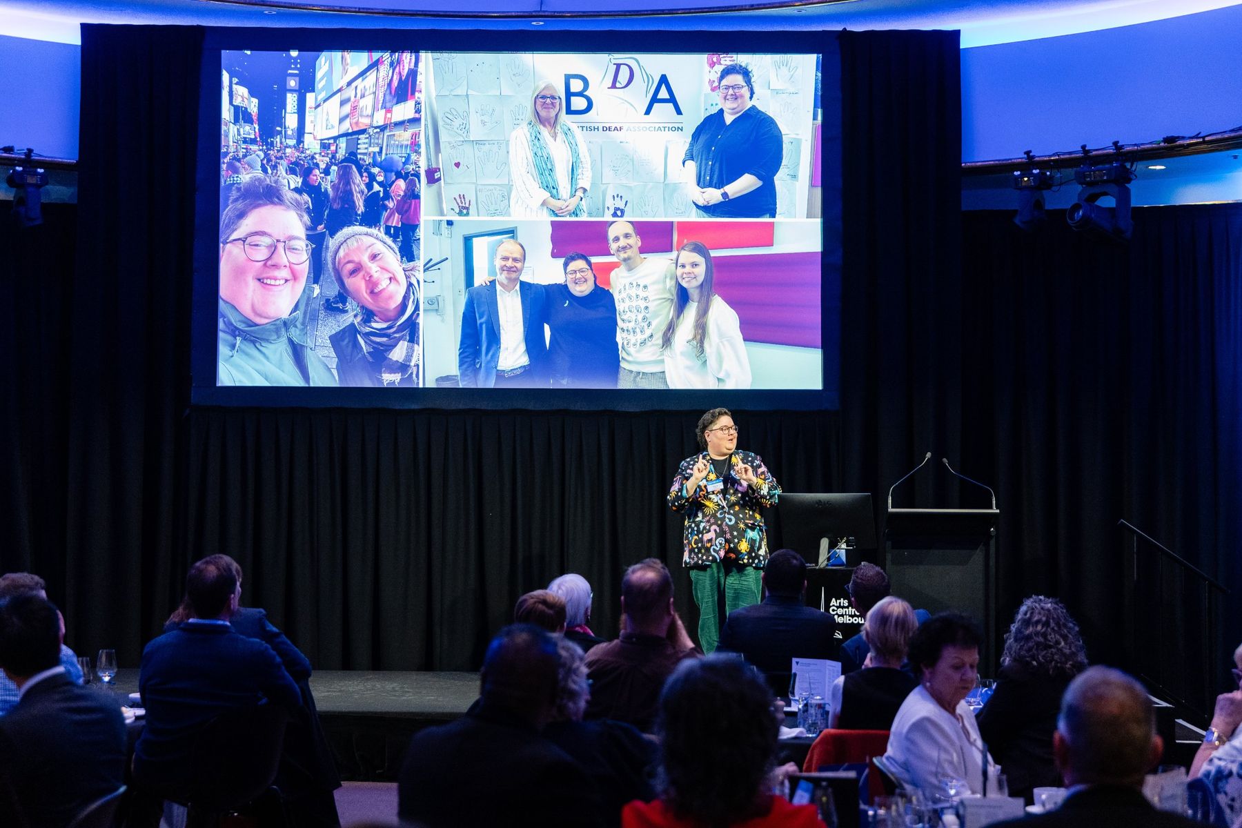 Sherrie signing on stage at Arts Centre Melbourne in front of a seated audience, with travel and Deaf community photos on the big screen behind.