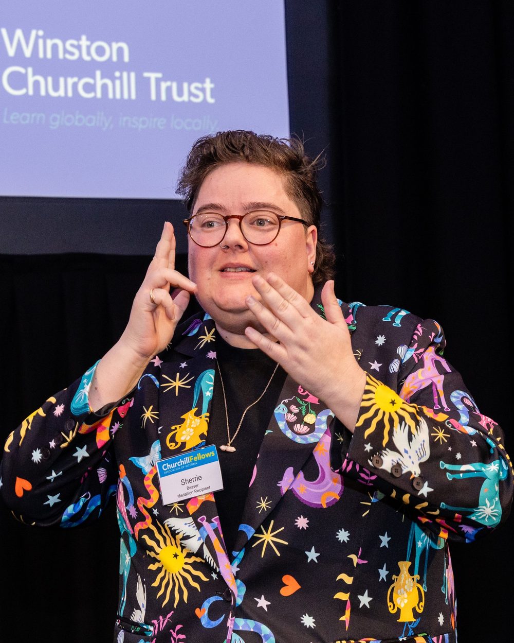 Sherrie signing in Auslan on stage at a Winston Churchill Trust event, wearing a bright patterned blazer and glasses.