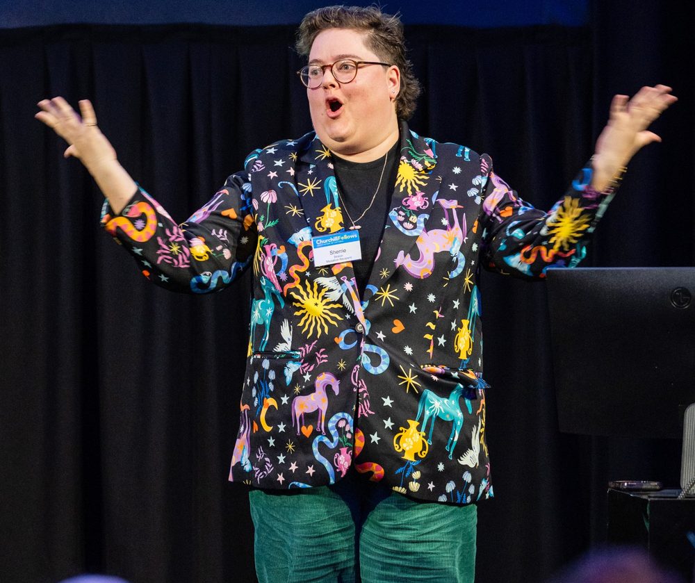 Sherrie on stage with arms raised and an excited expression, wearing a colourful patterned blazer.